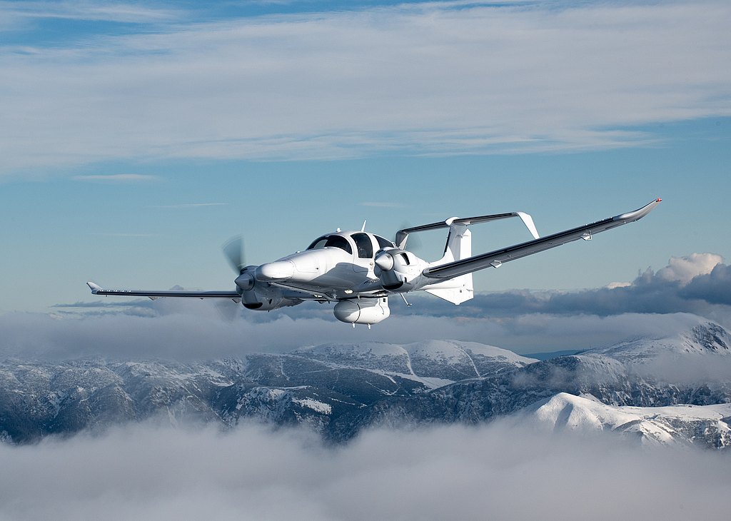 A sleek white aircraft soaring through a cloudy sky, with snow-capped mountains visible in the background.