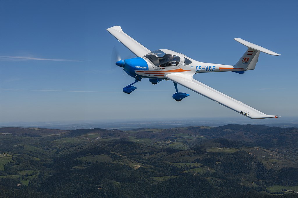 A blue and white DA20i Katana aircraft soars above lush green hills under a clear blue sky, showcasing its elegant design.