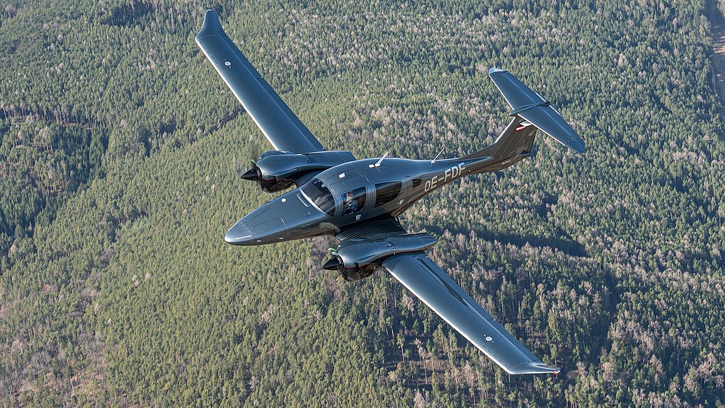 A DA62 aircraft glides through the sky, showcasing its shiny exterior against a backdrop of lush green forests below.