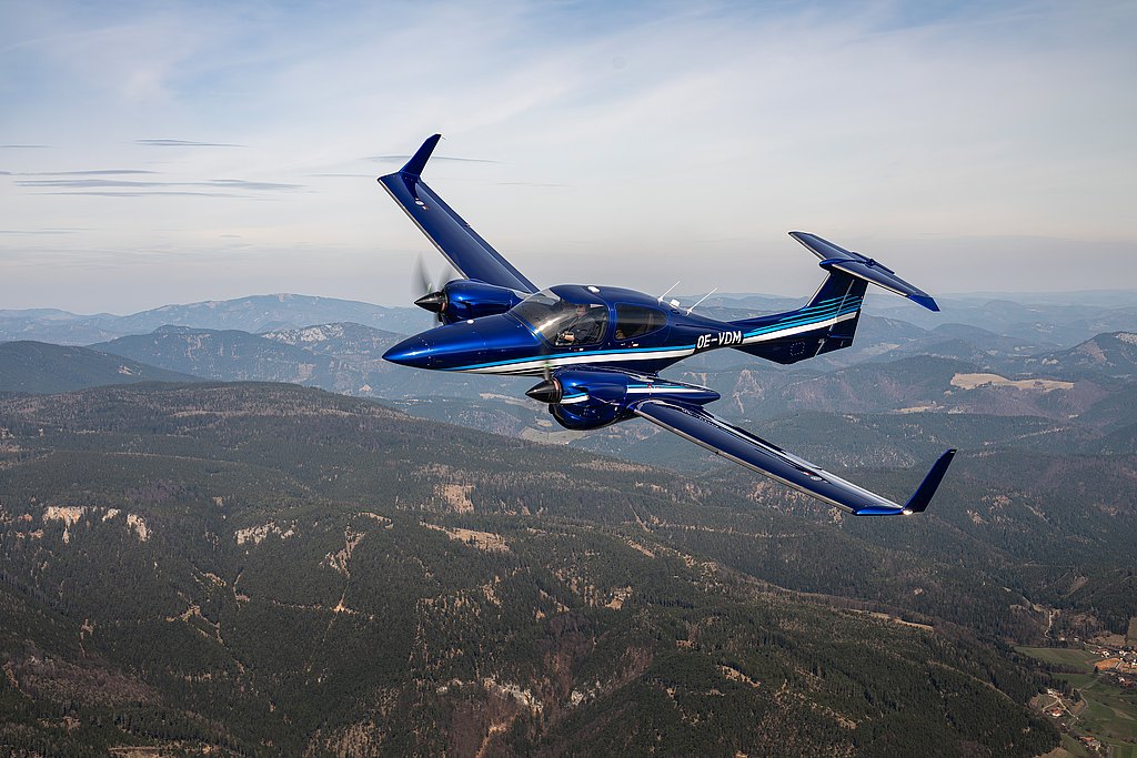 A blue DA42-VI aircraft soars above a lush green landscape, showcasing its elegant design against a backdrop of rolling hills.