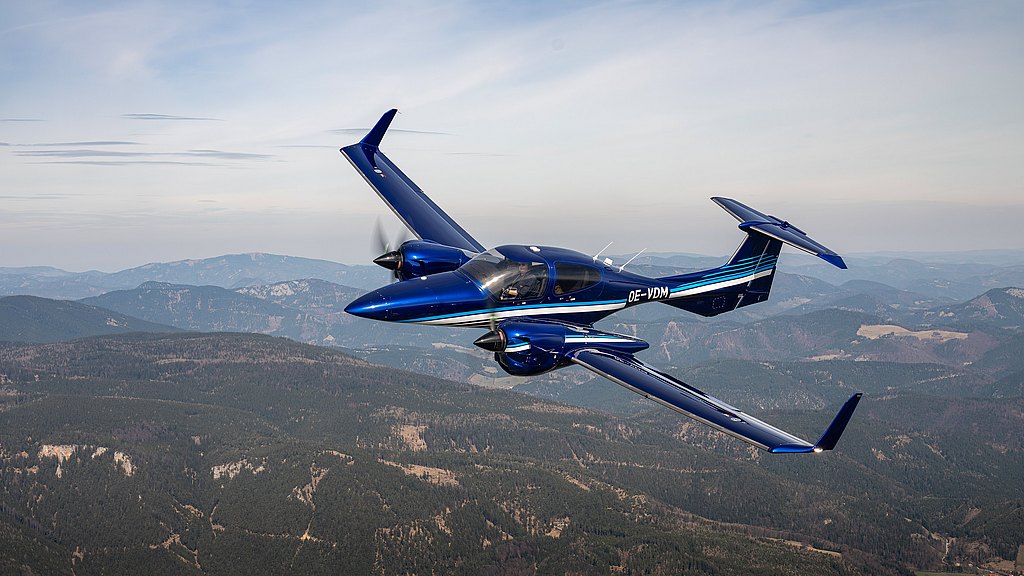 A blue DA42-VI aircraft soars above a lush green landscape, showcasing its elegant design against a backdrop of rolling hills.