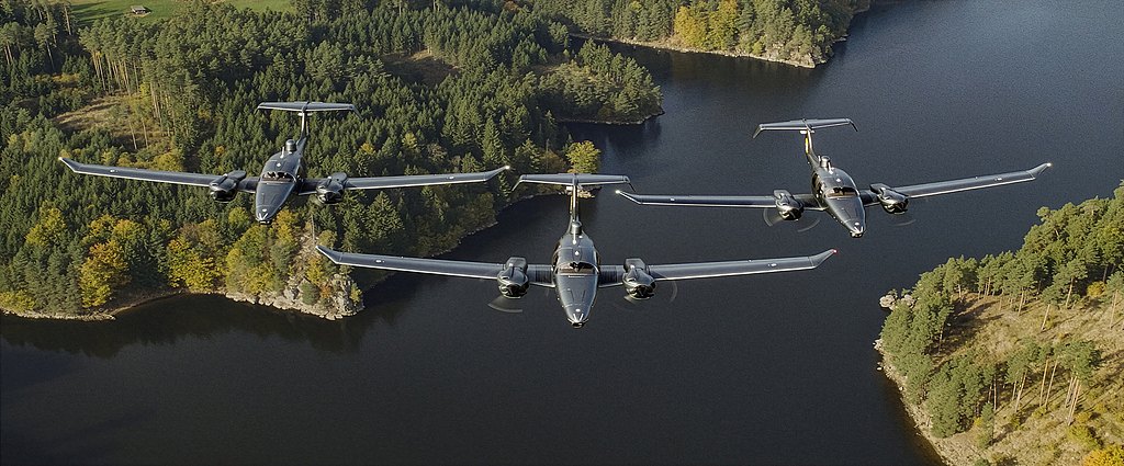 Three DA62 MPP aircraft fly in formation over a serene lake, surrounded by lush green forests and autumn-colored trees.