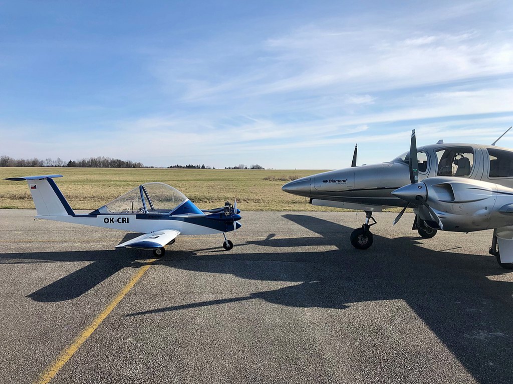 A small blue and white aircraft is parked in front of to a silver DA62 on a sunny day, with a grassy field in the background.