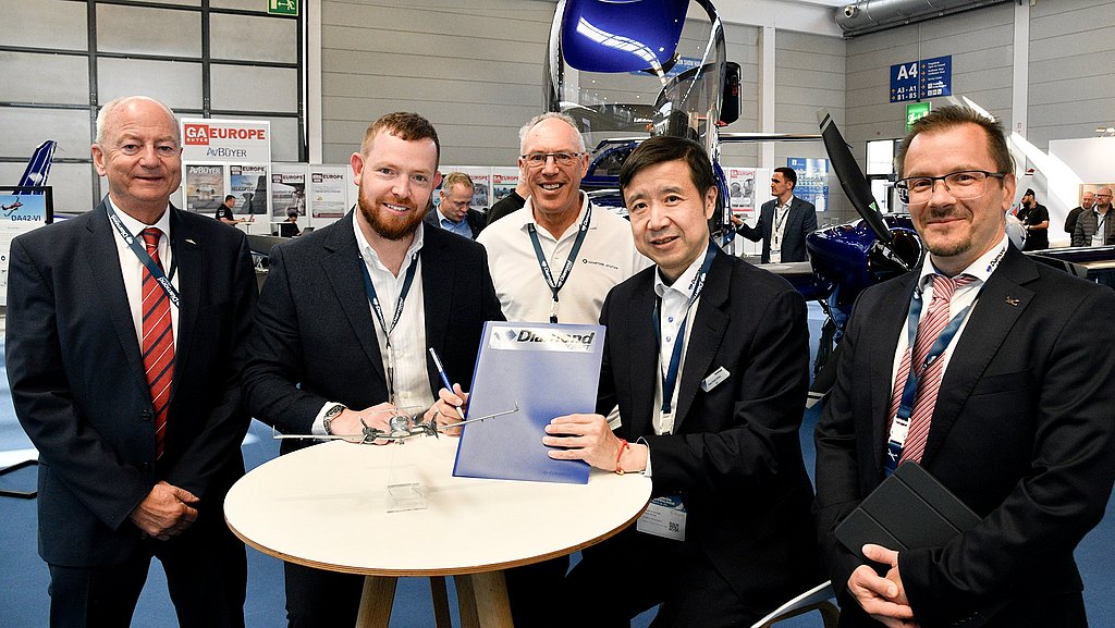 A group of four men stands around a table, celebrating a signed agreement, with a model aircraft displayed prominently.