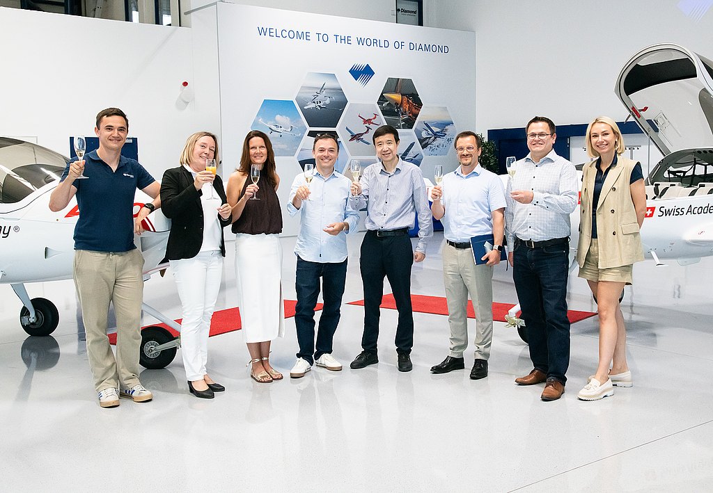 A group of eight people celebrate with drinks in hand, standing in front of a small aircraft in a bright, modern setting.