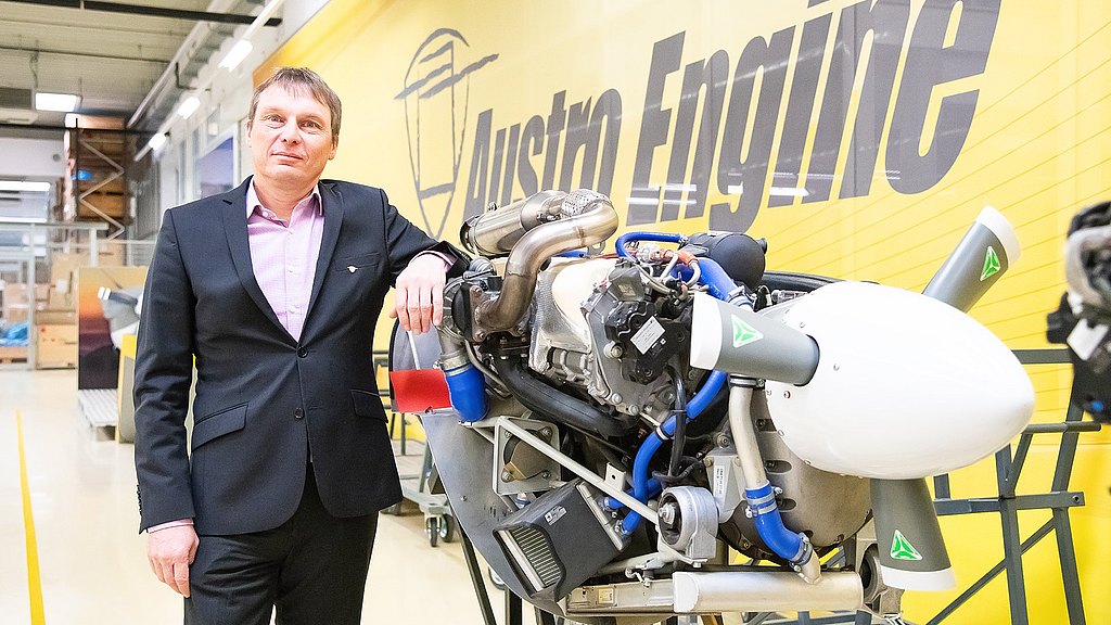 A man in a suit stands confidently beside a modern aircraft engine, showcasing advanced engineering in a bright workshop.