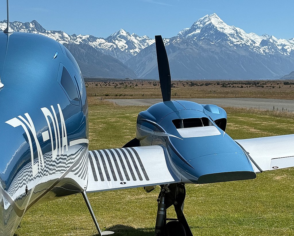 A sleek blue DA62 aircraft is parked on a grassy field, with majestic snow-capped mountains rising in the background under a clear sky.