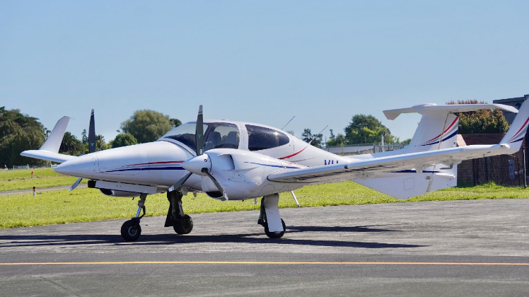 A sleek white DA42-VI aircraft with red and blue stripes is parked on a sunny airstrip, surrounded by green grass and trees.