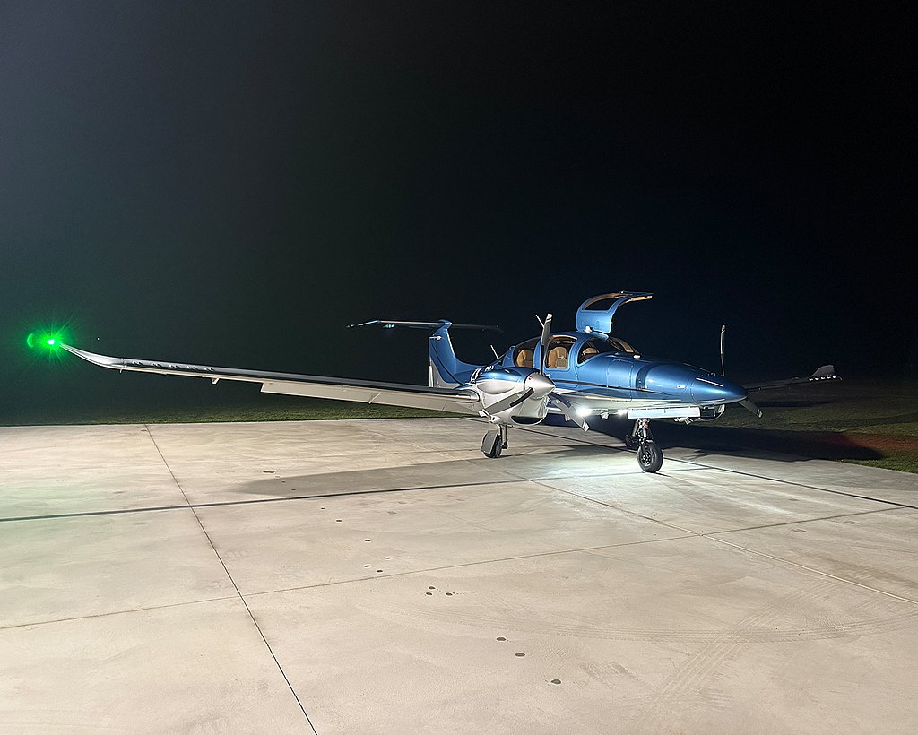 A blue DA62 aircraft with a green light on its wing is parked on the tarmac under a dark sky, getting ready for an early departure..