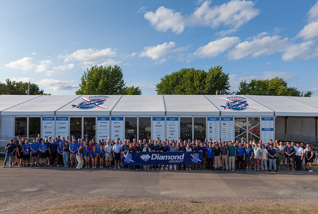 A large group of people stands together in front of a tent, proudly displaying a banner for Diamond Aircraft.