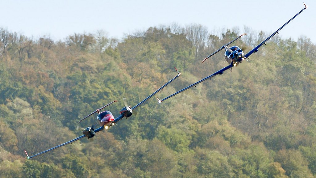 Blue DA50 RG and orange DA42-VI flying in formation with forest in the back.