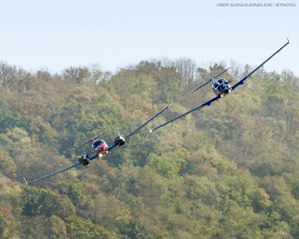 Blue DA50 RG and orange DA42-VI flying in formation with forest in the back.