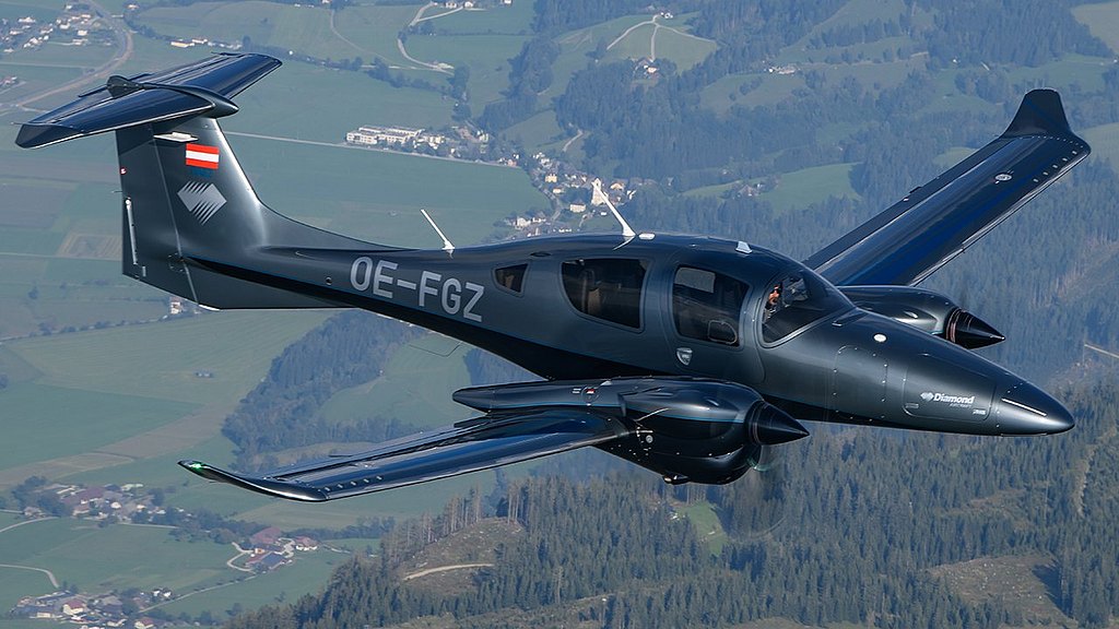 DA62 in anthracite and black design flying over a village and forest.