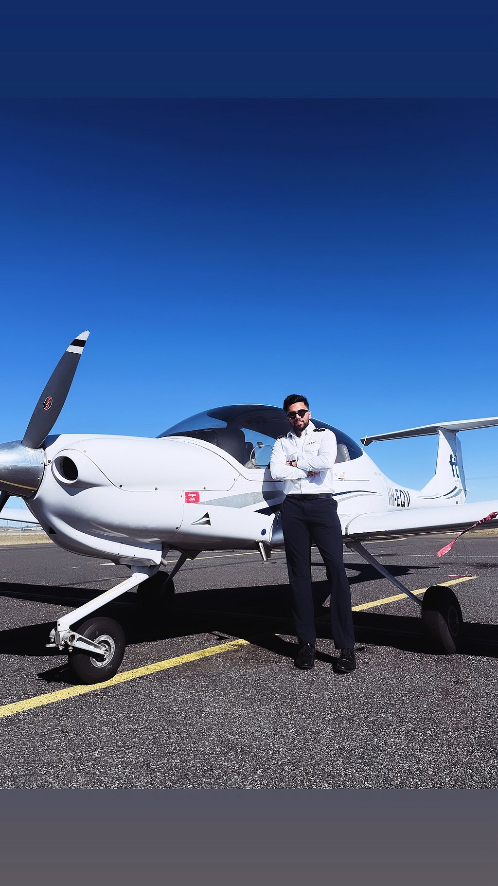 A confident pilot stands with crossed arms next to a sleek DA40 aircraft against a clear blue sky.