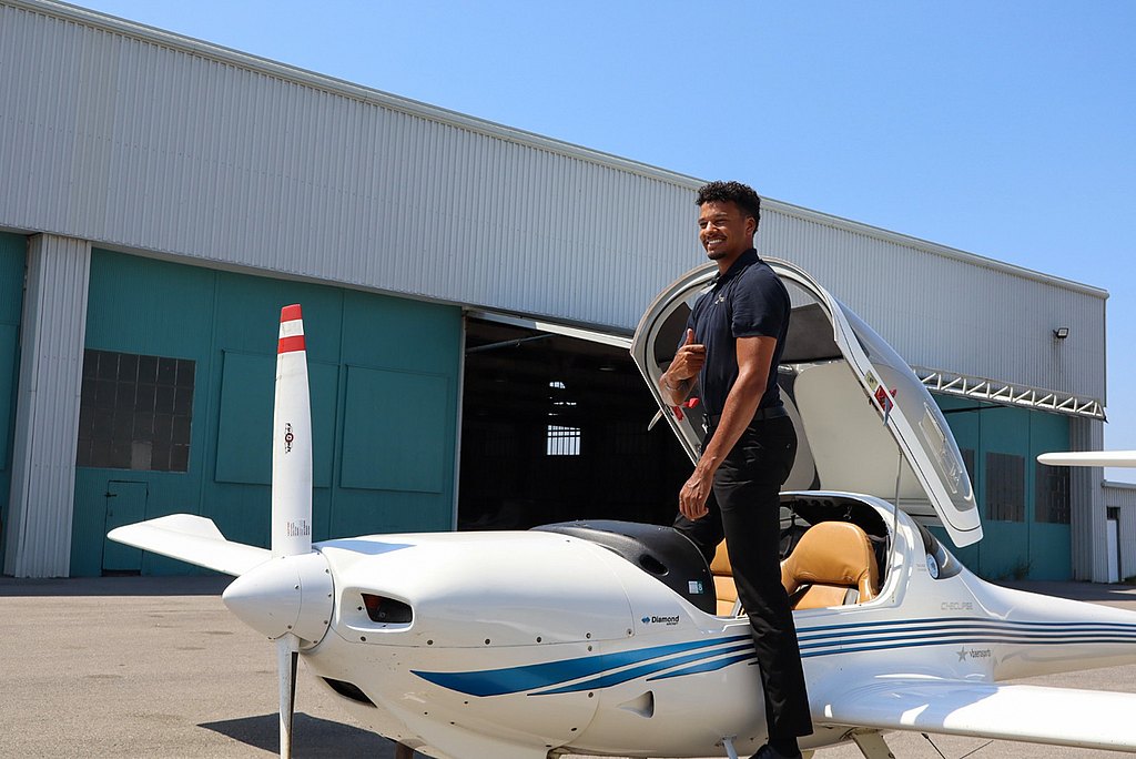 A young man stands on the wing of a DA20 aircraft, smiling and giving a thumbs-up, with a hangar in the background.