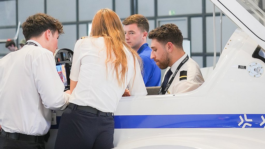 A group of student pilots gathers around a DA20 aircraft, focused on the controls and screens in front of them.