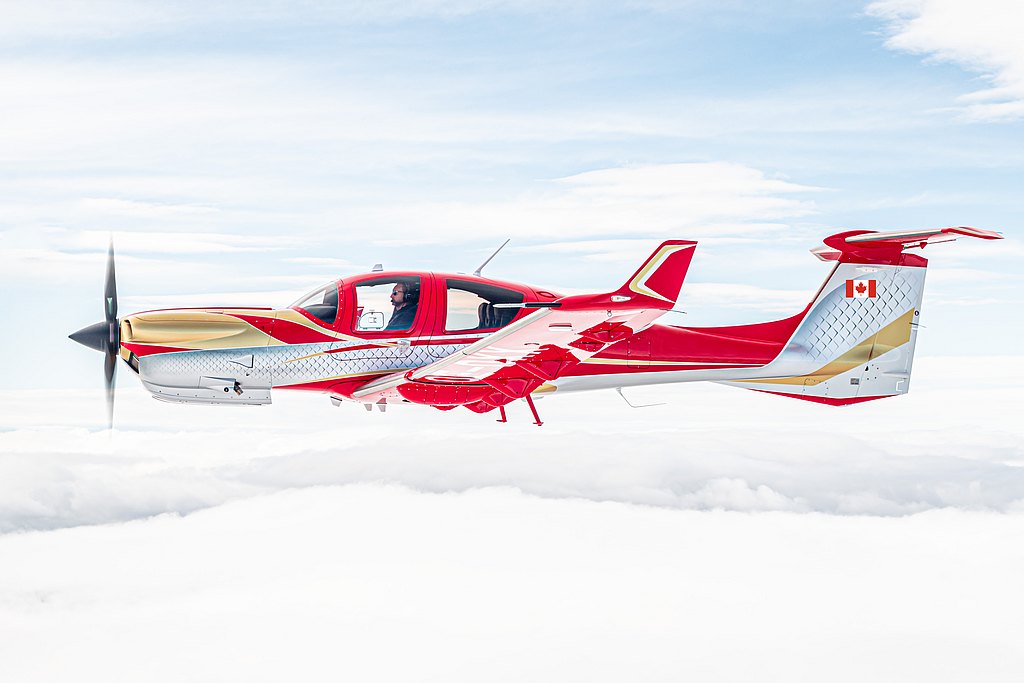 The vibrant DA50 RG aircraft, adorned with a Canadian flag, glides gracefully through a bright sky, surrounded by fluffy white clouds.