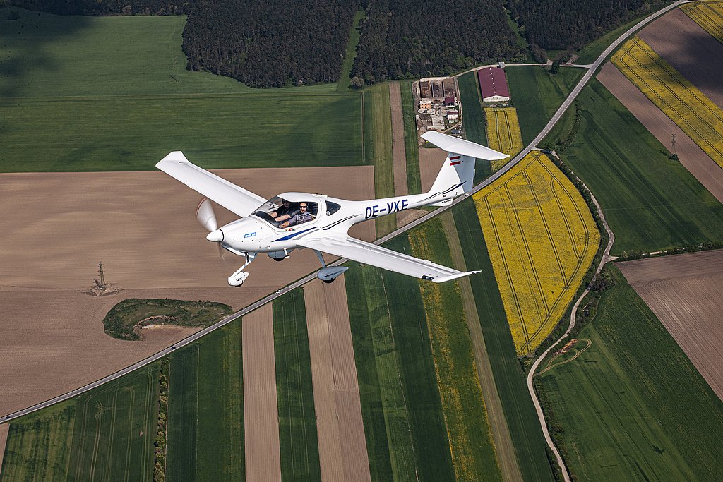 A DA20i Katana aircraft flies over vibrant green and yellow fields, showcasing the beauty of rural landscapes from above.