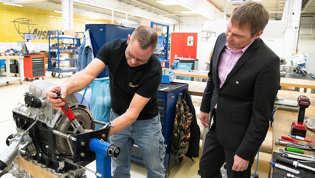 Man standing next to a production working, who is working on an engine.