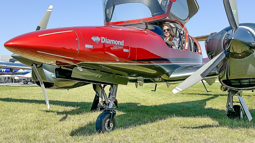 Female pilot sitting in the pilot seat of a orange DA42-VI standing on grass. 