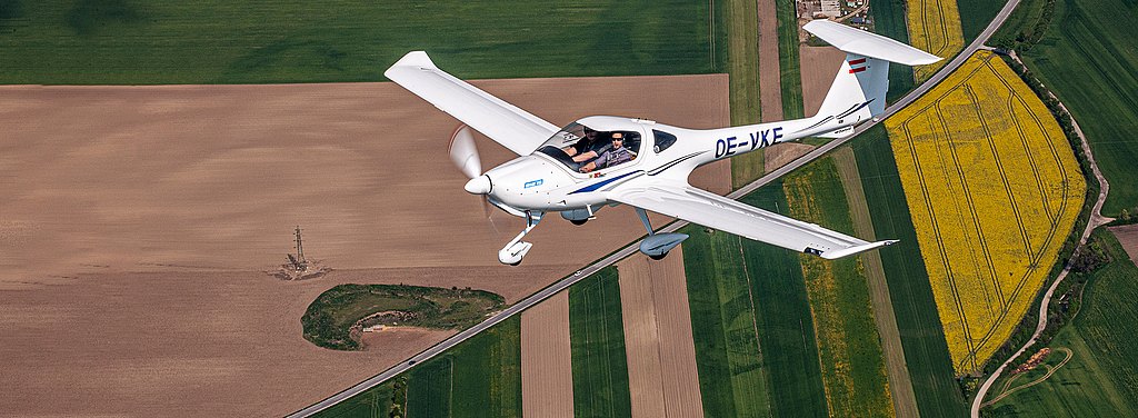 Two pilots navigate a sleek DA20 aircraft, gliding over a colorful patchwork of farmland, with bright yellow flowers contrasting against green fields.