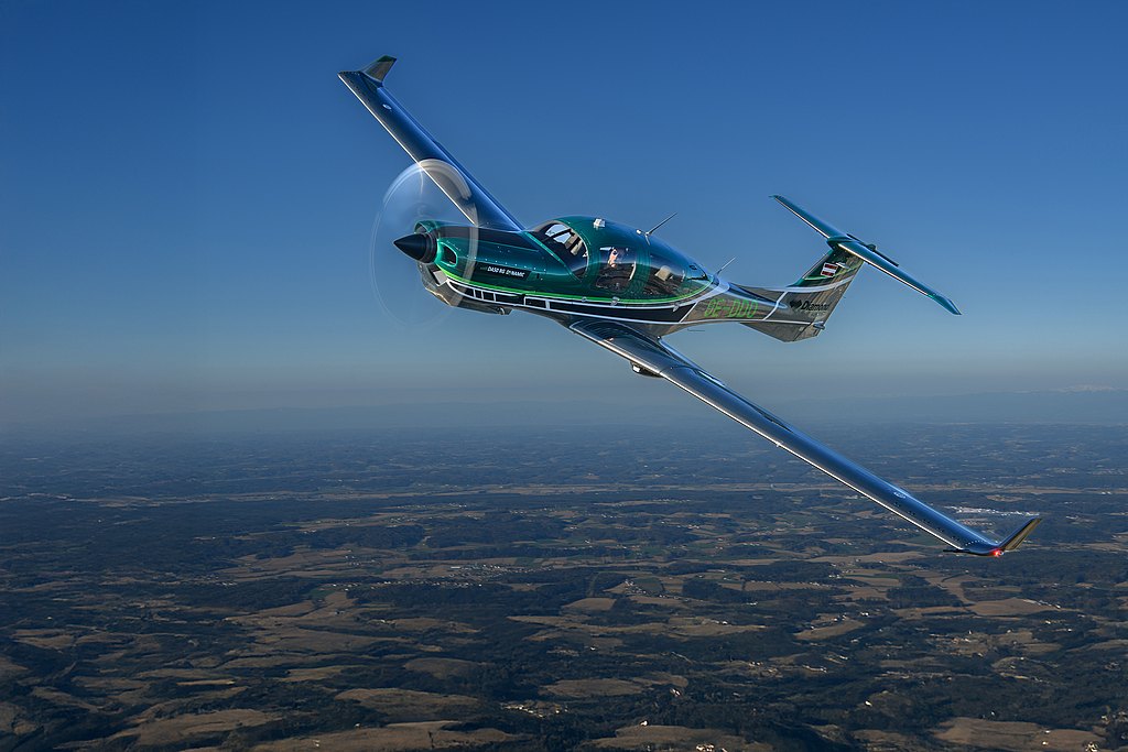 A DA50 RG aircraft soars through a clear blue sky, showcasing its shiny wings and propeller against a scenic landscape below.