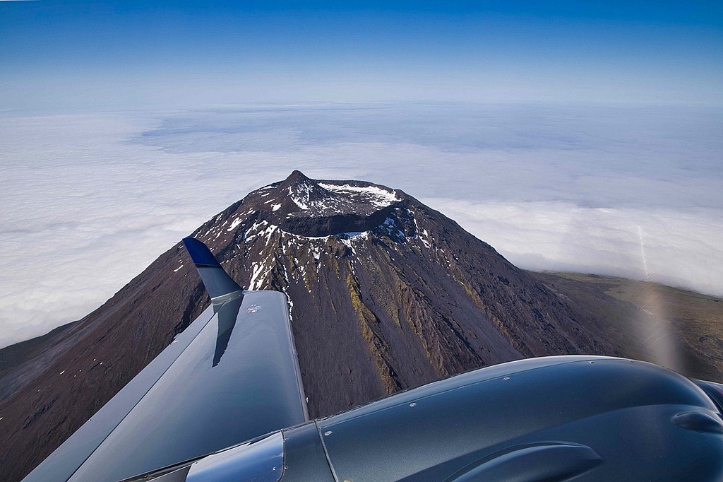 Aerial view of a snow-capped volcano peak rising above a sea of clouds, with an airplane wing in the foreground.