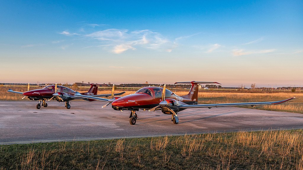 Two red and silver DA62 aircraft are parked on a tarmac, surrounded by tall grass under a clear blue sky.