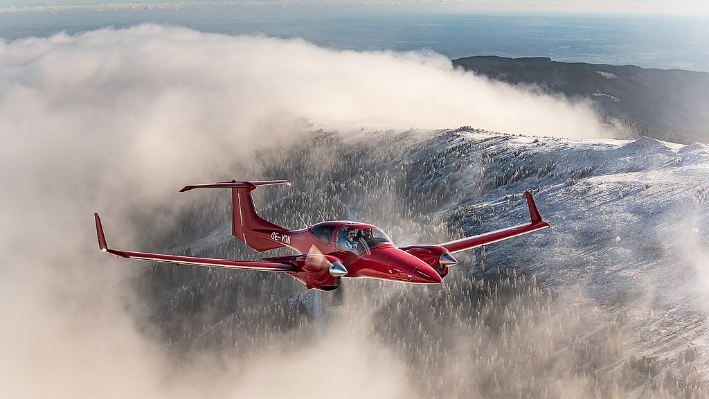 Red DA42-VI flying over snowy, foggy mountain.