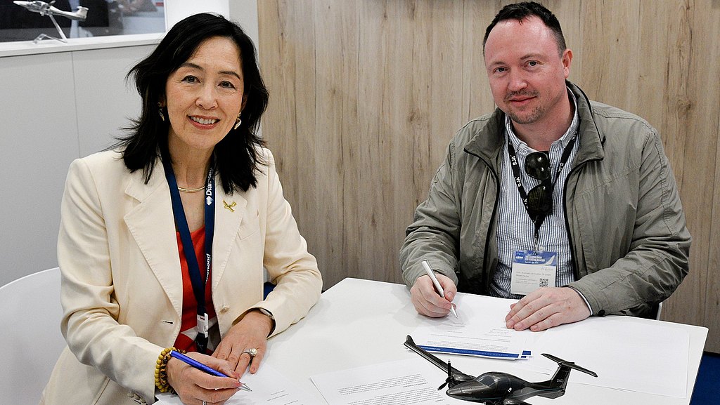 A woman in a cream blazer smiles while signing documents at a table, with a model airplane in front of her.