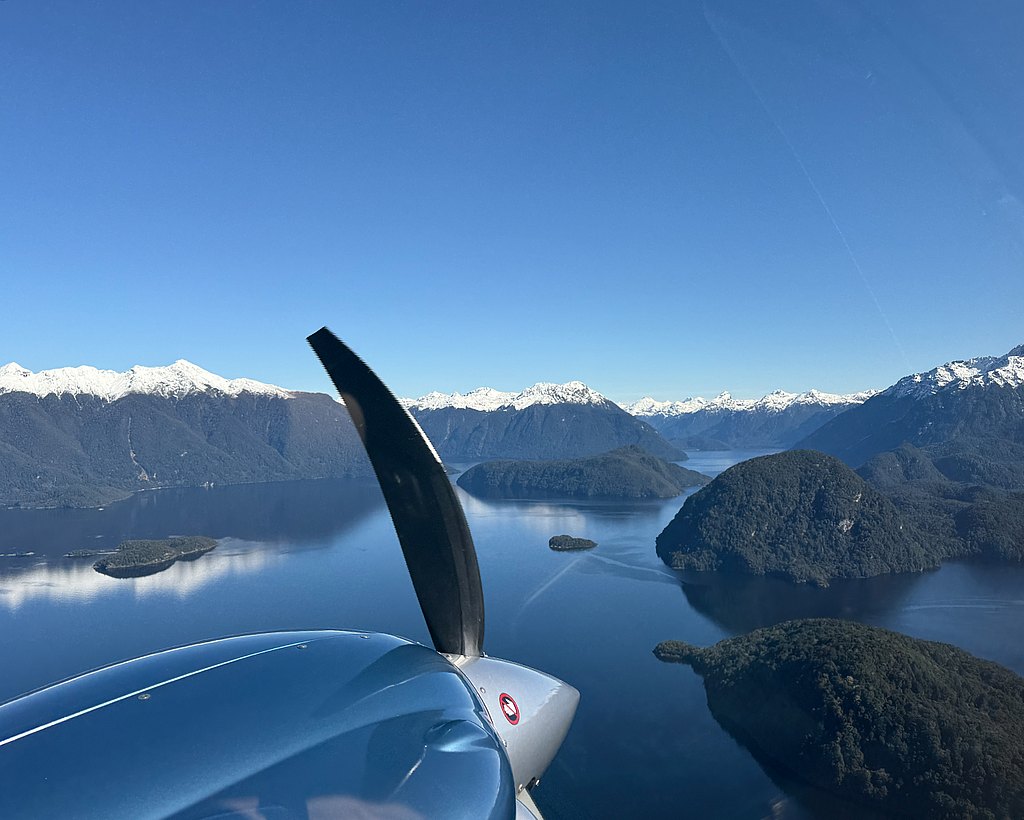 Aerial view from a DA62 pilot seat on a  lake surrounded by lush green islands and majestic snow-capped mountains under a clear blue sky.