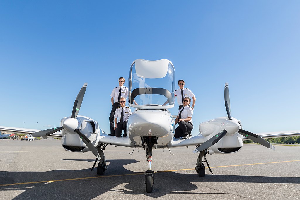 Four student pilots pose confidently on a DA42 aircraft, showcasing their uniforms against a clear blue sky at an airport.