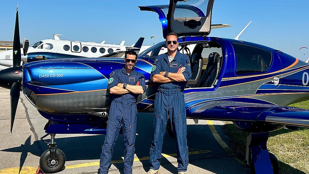 Two Diamond Aircraft pilots in flight suits standing in front of a blue DA50 RG with open gullwing doors. 