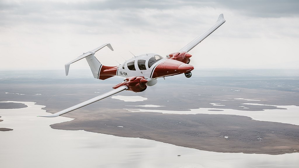 DA62 MPP in a red an white design flying over a lake.