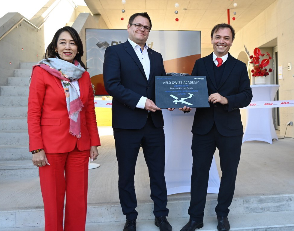 In a modern setting, a woman in red and two men in suits proudly present a plaque for the Aelo Swiss Academy.