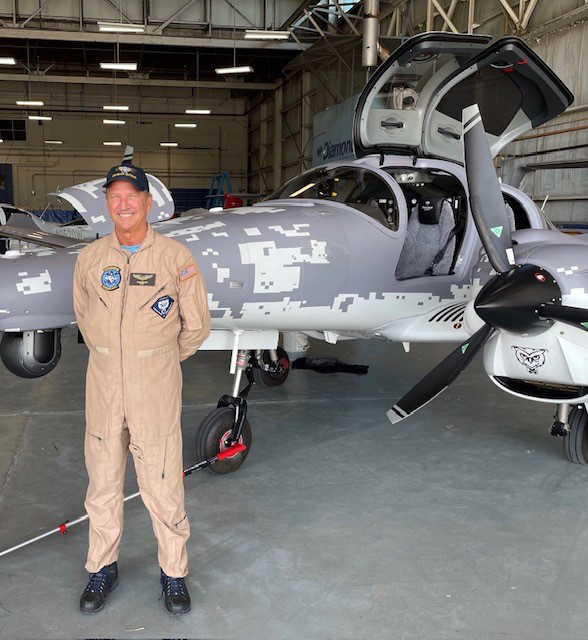 A smiling pilot in a beige flight suit stands proudly next to a gray DA62 MPP aircraft with a unique camouflage pattern.
